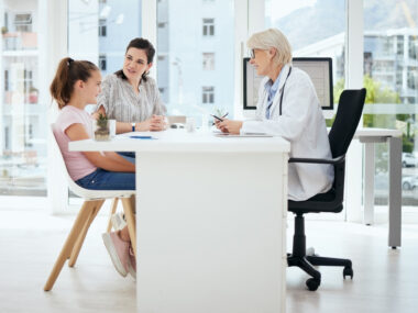 A doctor sits at a desk across from a young patient and their parent.
