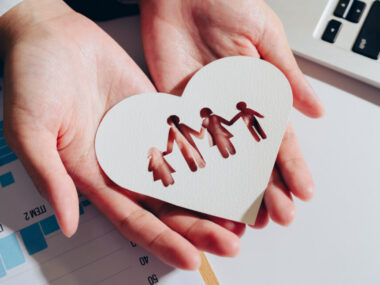 A pair of hands hold a heart-shaped piece of paper with cutouts of figures representing family and symbolizing love and care.